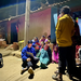 Several children and others sit and stand around Eshaan Patheria who has a laptop on his lap. The scene is illuminated by a street lamp, and a lightbulb is on in the house behind them.