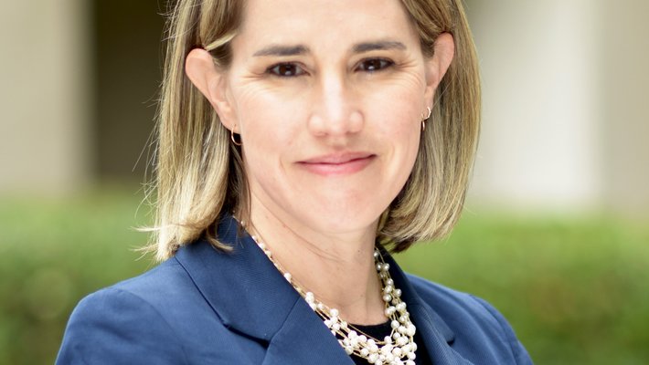 A photo portrait of a woman with blond hair wearing a blue blazer and black shirt with a chunky pearl necklace.
